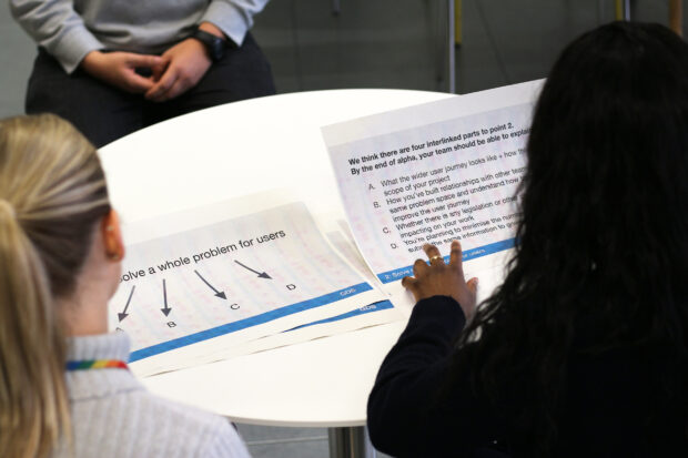 An assessor forum: Two people sitting at a table with two documents in front of them reading “Solve a whole problem for users” and “We think there are four interlinked parts to point 2. By the end of alpha, your team should be able to explain…”; another person is sitting opposite of them in a hot chair, responding to their questions