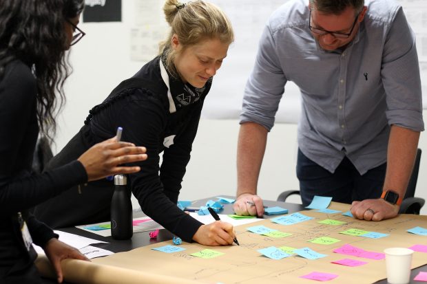 Three people in a workshop creating a user journey map on a brown paper roll with coloured sticky notes