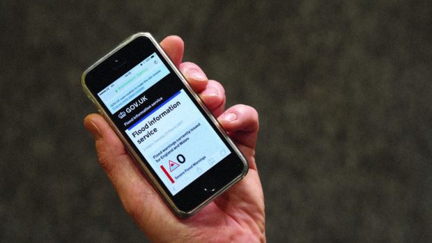 A smartphone in person's hand, displaying the flood information service page on GOV.UK, indicating the current flood warnings in England and Wales.