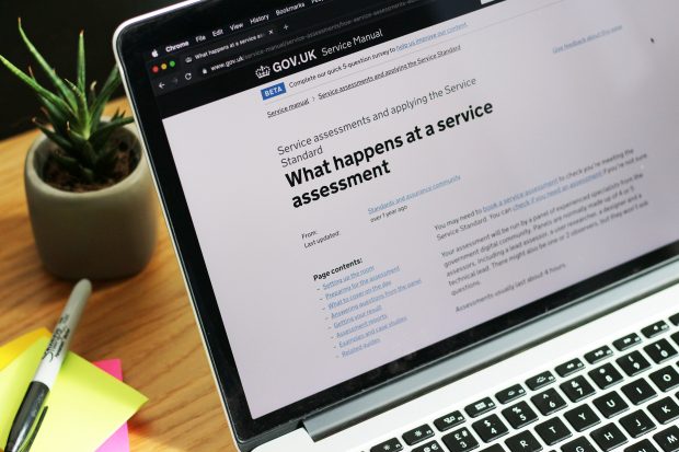 A laptop on a desk next to a pile of fresh sticky-notes; its screen shows guidance from the GOV.UK Service Manual on ‘What happens at a service assessment’ from the ‘Service assessments and applying the Service Standard’ section