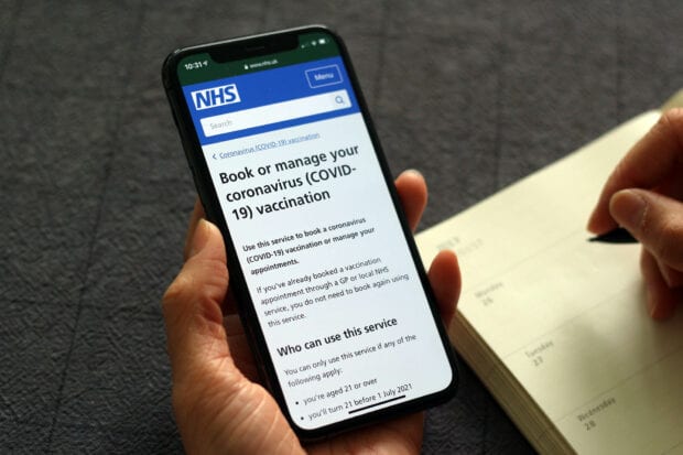 The NHS Book your coronavirus vaccination online service shown on a mobile phone held by a person who’s taking a note in their diary