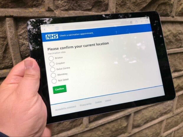 A person’s hand holding a tablet computer showing the web application for checking a vaccination appointment used by NHS staff in vaccination centres