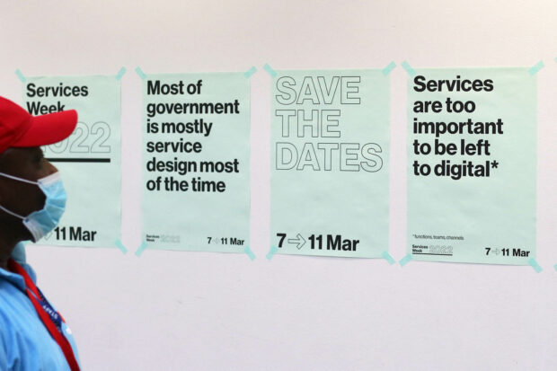 A series of posters announcing Services Week 2022 on a wall, a man looking at them