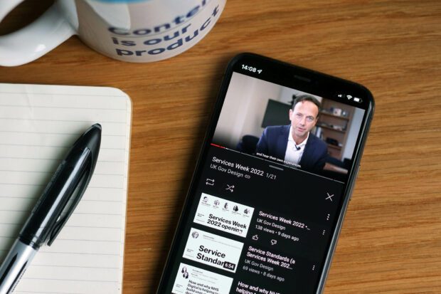 A mobile phone showing a YouTube playlist of recorded Services Week 2022 sessions with the welcoming note from Alex Chisholm, Chief Operating Officer for the Civil Service, playing