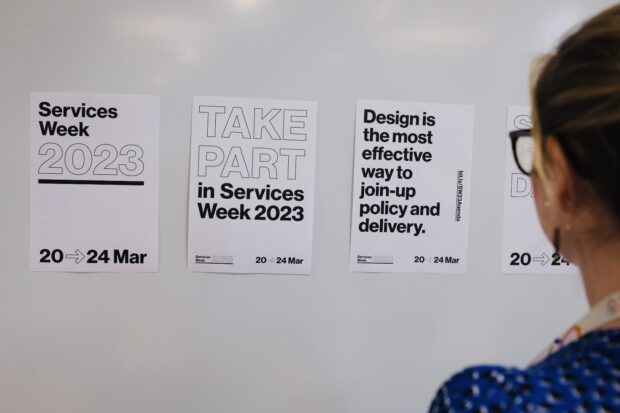 A woman wearing glasses looks at 3 Services Week posters on a wall. The posters read: Services Week 2023. Take Part in Services Week 2023. Design is the best way to join up policy and delivery. All have the date on them: 20 to 24 Mar.