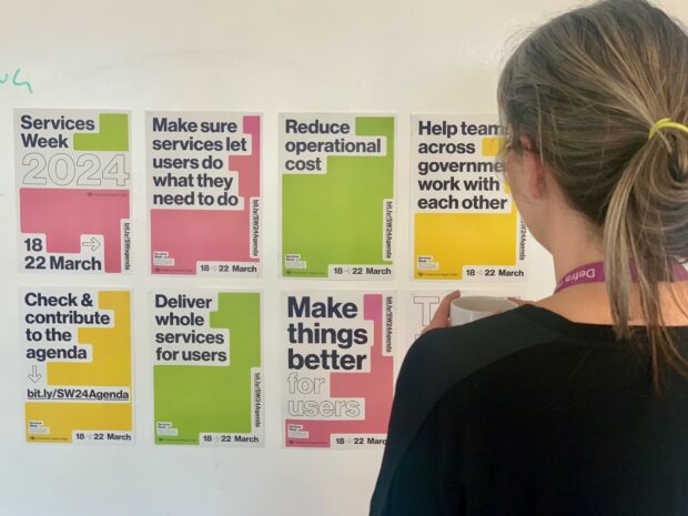 A person with a ponytail and a cup of drink looks at a colourful series of Services Week 2024 posters on wall.